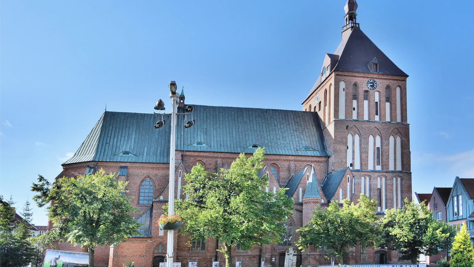 Katedra Niepokalanego Poczęcia NMP w Koszalinie, ceglana wieża z zegarem i zielony dach nad drzewami, błękitne niebo